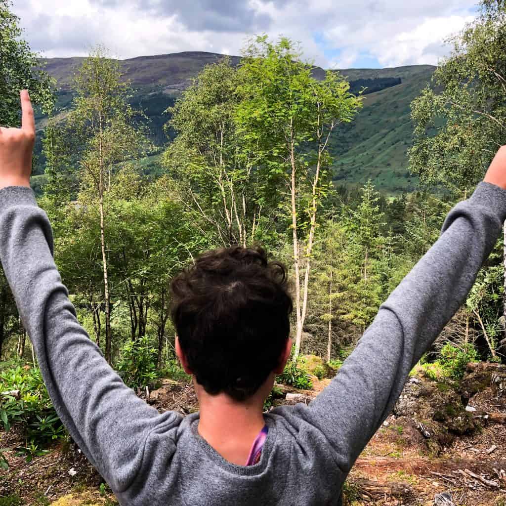 Boy with arms raised looking out over a Scottish valley
