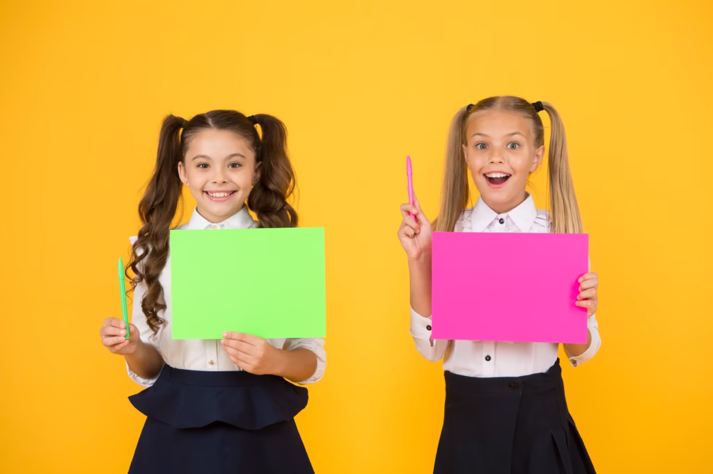 Two girls against a yellow background smiling and holding pink and green pieces of card up